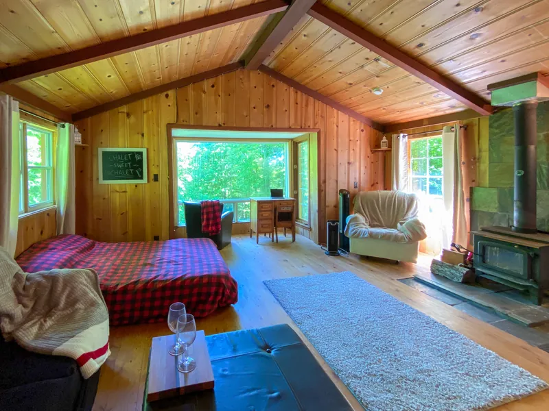 Pièce principale aux murs et plafond en pin, avec un poêle à bois et une grande fenêtre sur la forêt