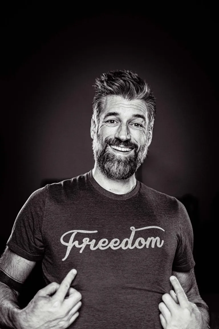 Portrait en noir et blanc d'Alexandre Comtois, souriant, qui pointe son t-shirt des deux mains